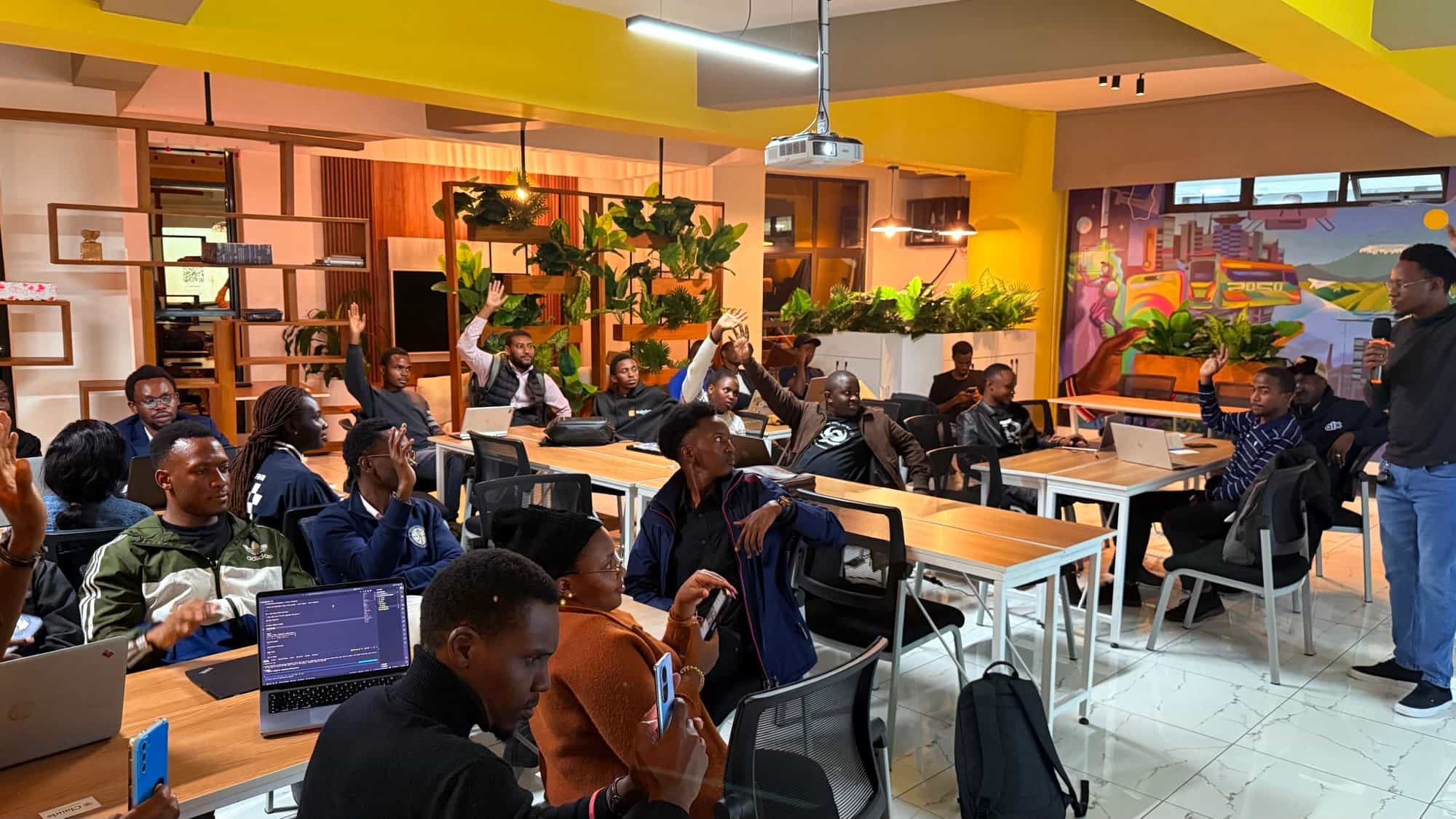 Workshop attendees raising their hands to answer a question, with a presenter speaking at the front of a bright, colorfully muraled room.