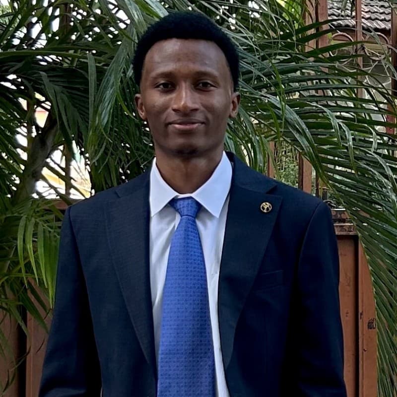 Billy Mwangi in a navy suit and blue tie, standing in front of palm leaves.