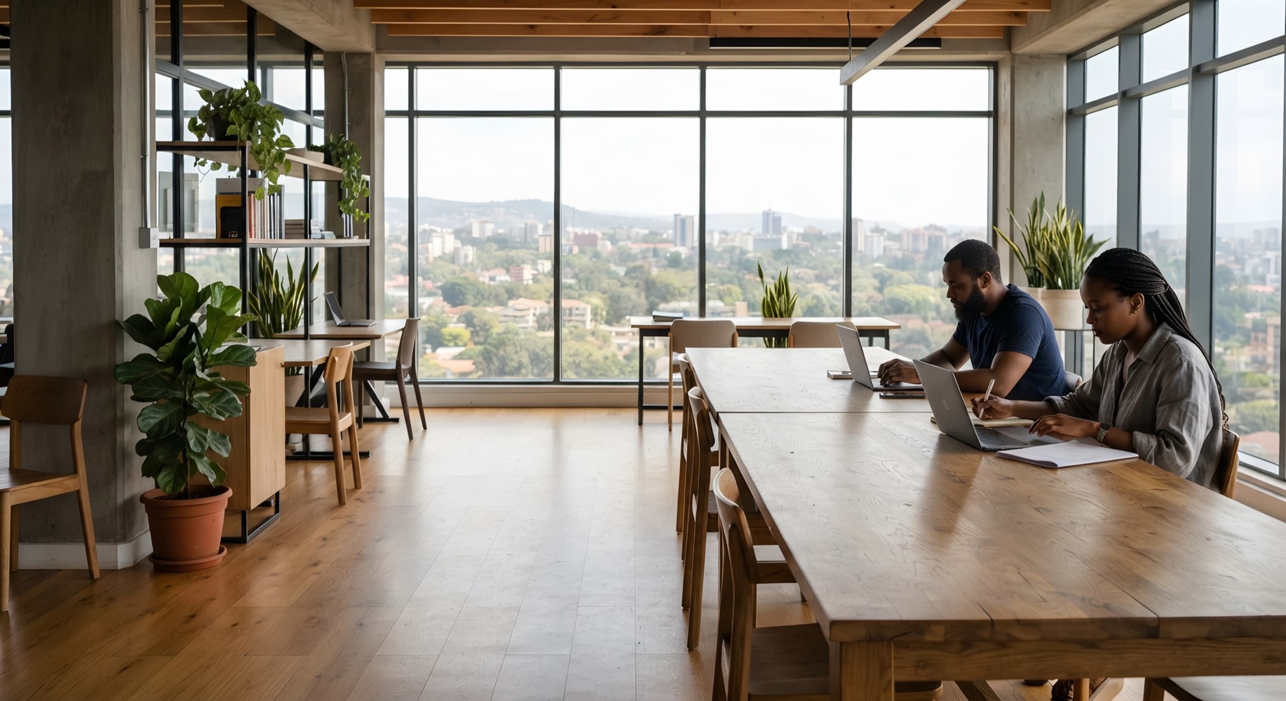 Two people work on laptops at a long wooden table in a bright office with windows over Nairobi.