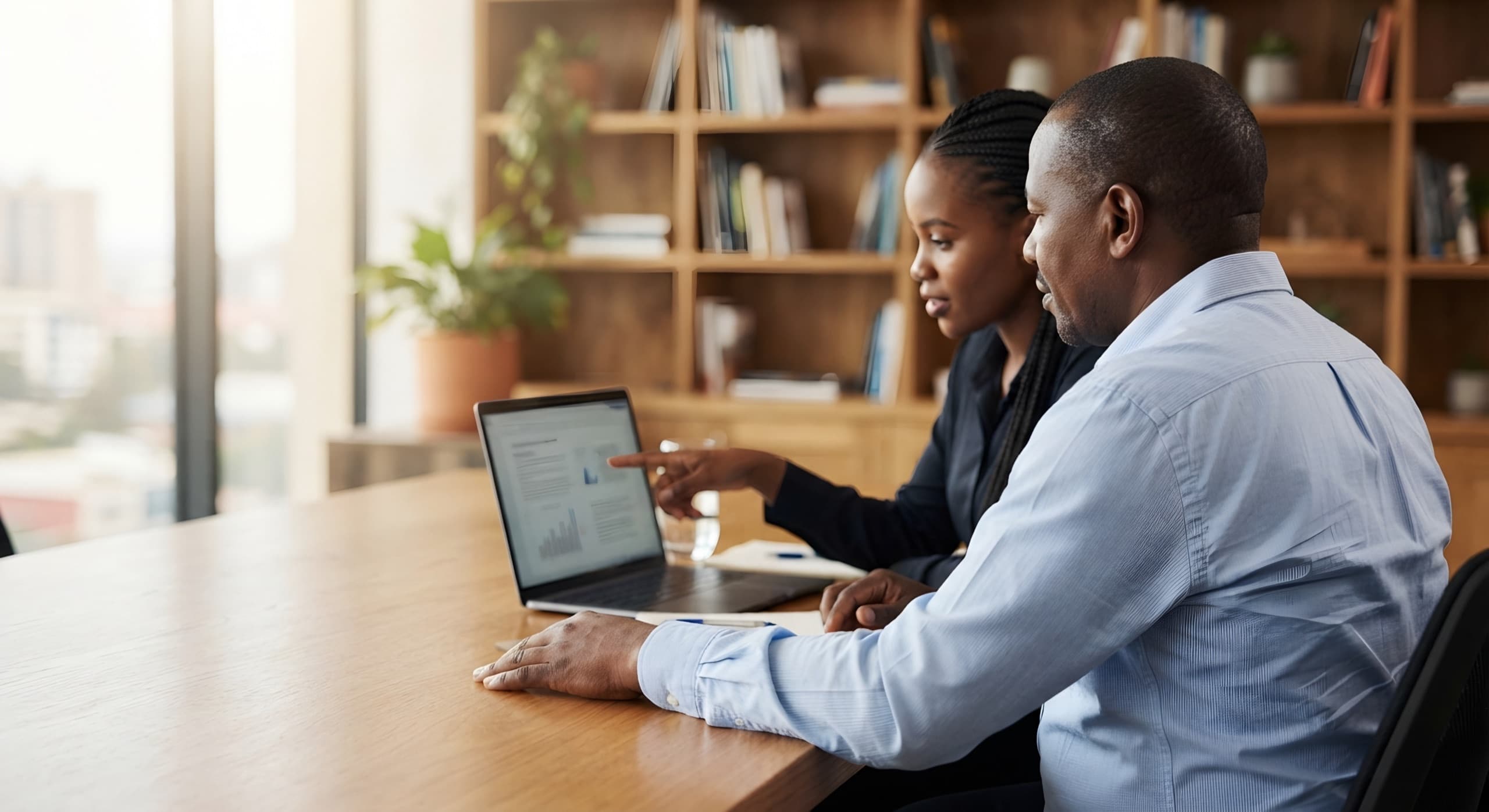 A man and a woman work side by side at a laptop in a quiet office lined with bookshelves.